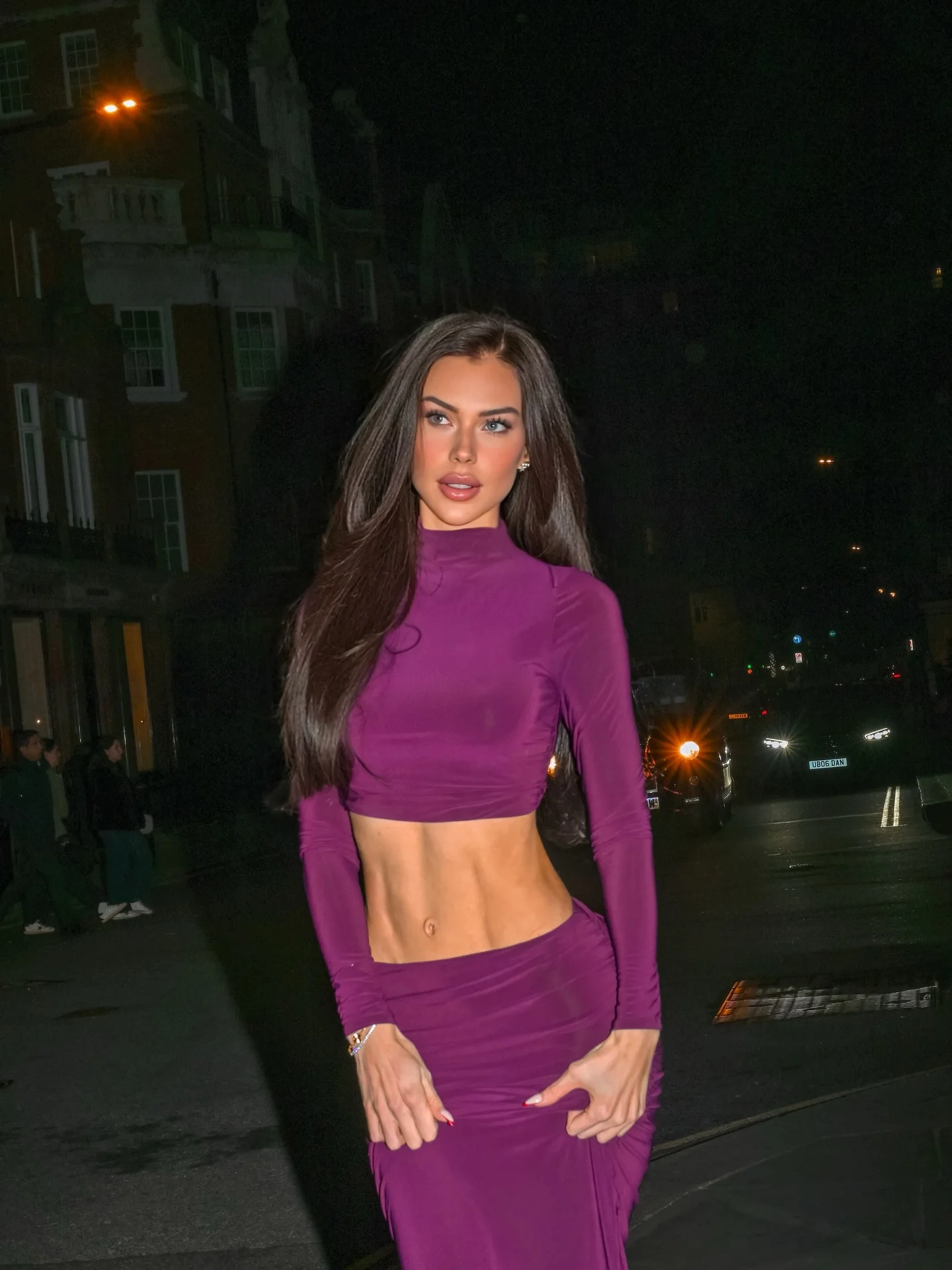 Brunette woman in a purple two-piece outfit beside a London street at night