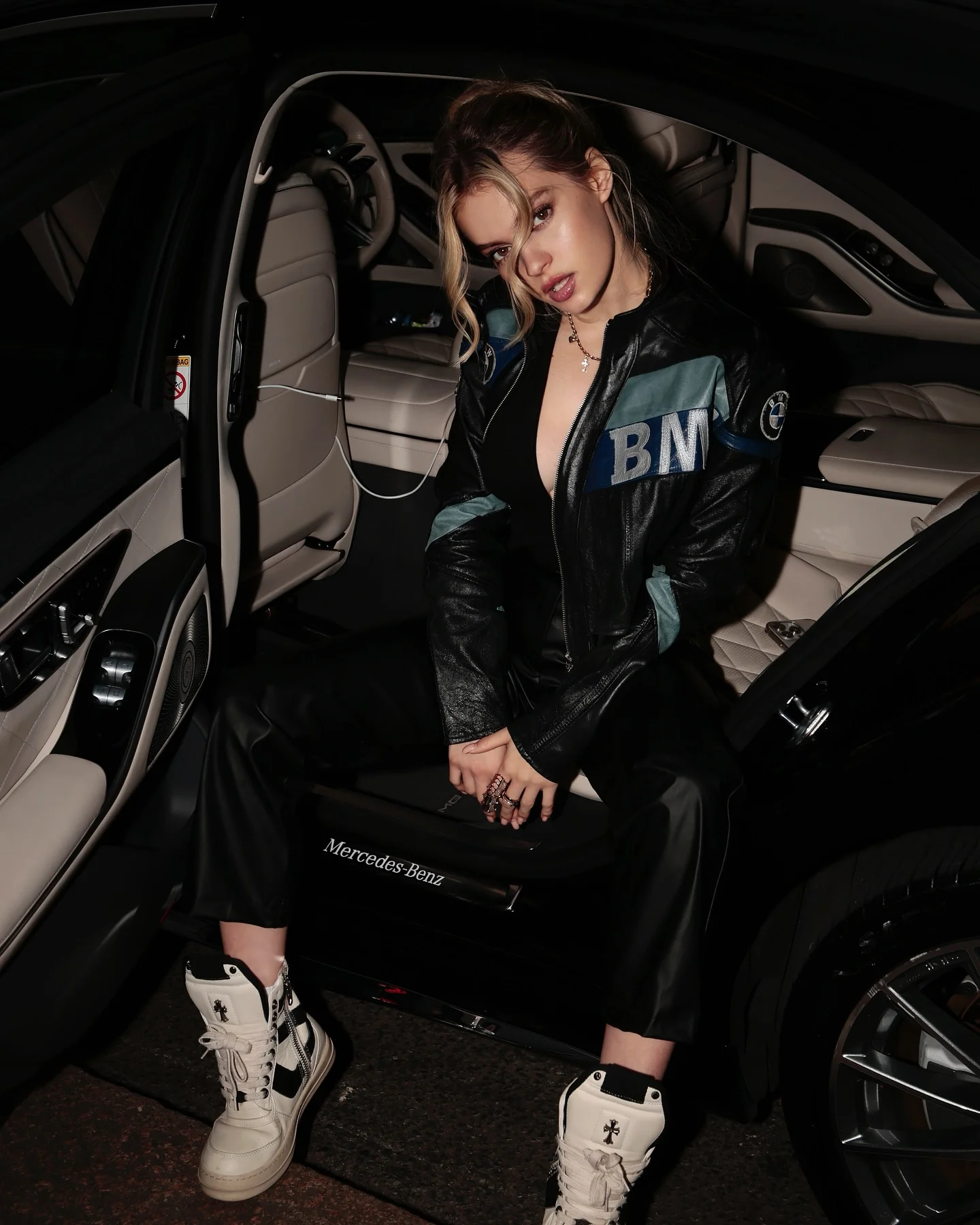 Woman in a black jacket and white trainers seated in an open car doorway at night