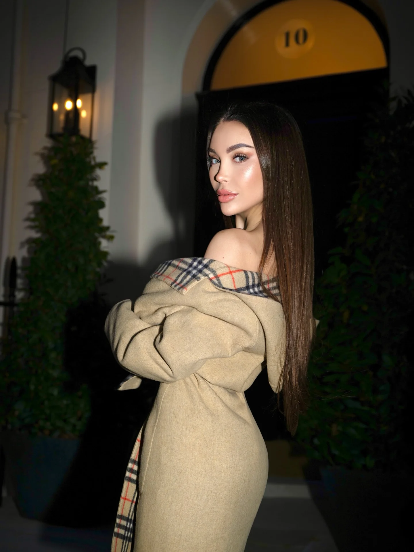Woman in a beige trench coat looking back beside a lit doorway