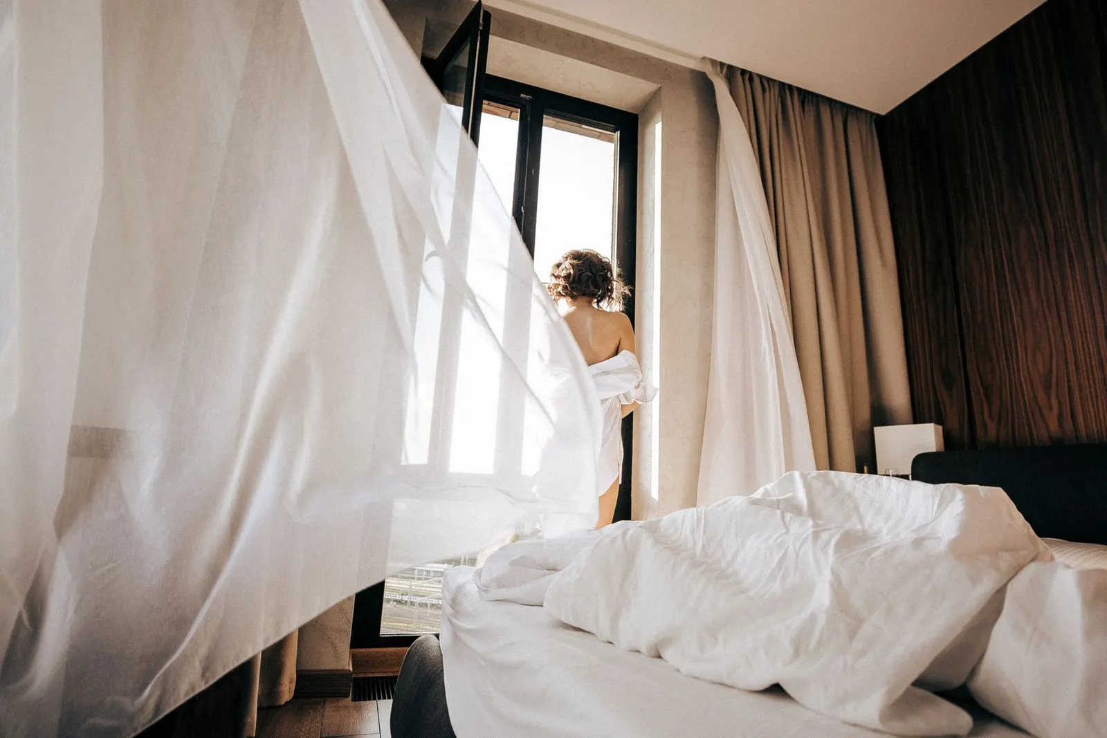Hotel room with a curtain blowing and a woman wrapped in a sheet at the window