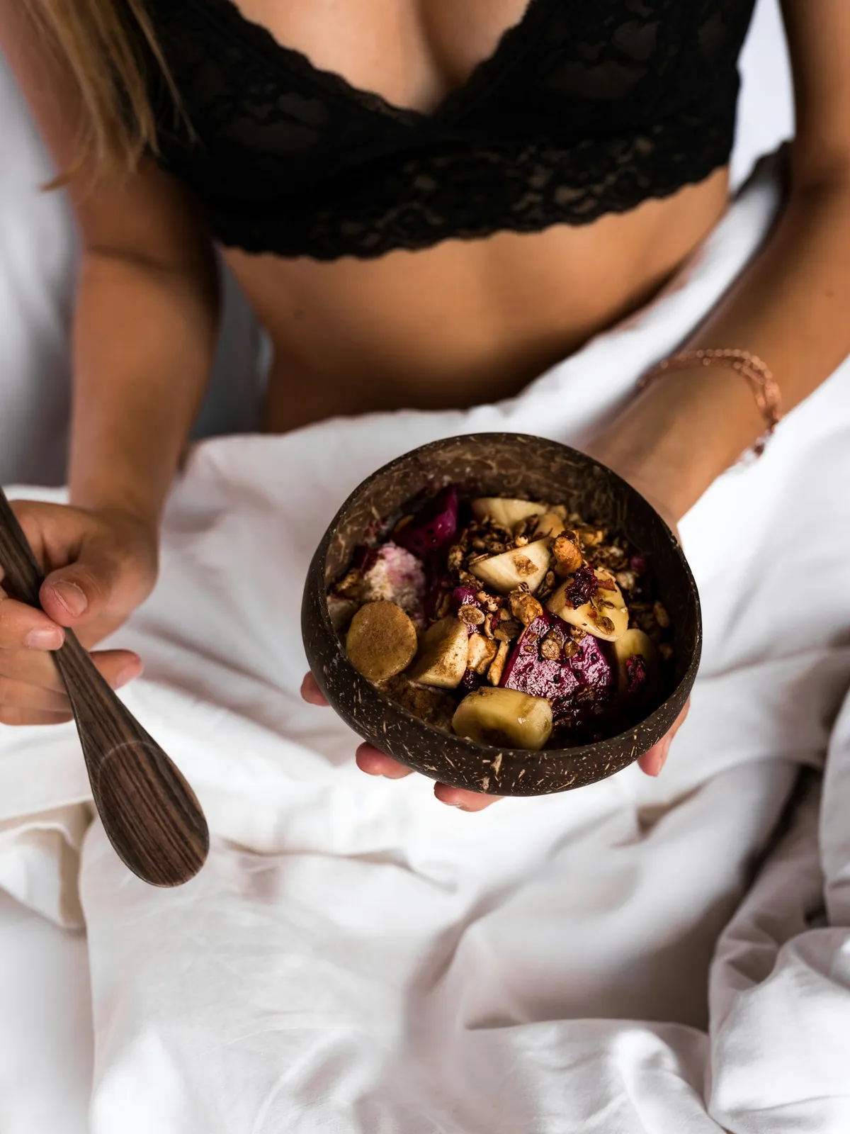 Woman in a black lace bra holding a fruit bowl in bed, her face out of frame