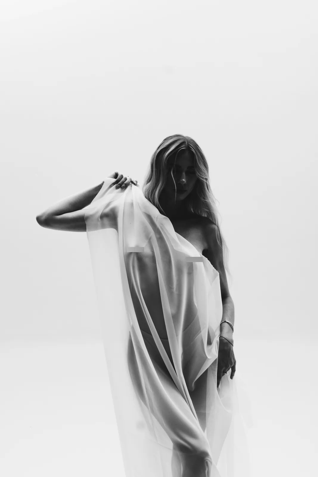 Woman draped in sheer white fabric against a white wall, in black and white